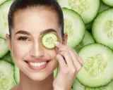 Una mujer sonríe mientras se coloca una rodaja de pepino en el ojo, rodeada de más rodajas de pepino.