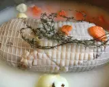 Préparation de la cuisson tête de veau cocotte. Le veau est ficelé, entouré de carottes et de thym dans un bouillon.
