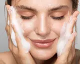 Donna con gli occhi chiusi mentre si lava il viso con un detergente schiumoso. Un gesto delicato per capire come si usa il detergente viso.