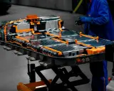 Assemblaggio di batterie per macchine elettriche, con cavi arancioni e celle metalliche. Un tecnico in tuta blu lavora sul pacco batteria.