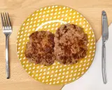 Dos hamburguesas para bebes, doradas y jugosas, servidas en un plato amarillo con puntos blancos. Cubiertos a los lados.