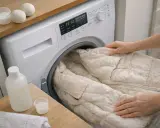 Ręce wkładają beżową kurtkę puchową do pralki. Obok butelka płynu i kulki do prania.