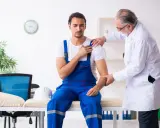 Un ouvrier en bleu de travail consulte un médecin pour une possible maladie professionnelle de l'épaule. Le médecin examine son bras.
