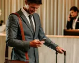 Biznesmen z walizką sprawdza telefon, być może szukając informacji o tym, co to jest OTA, podczas gdy recepcjonistka rozmawia przez telefon.