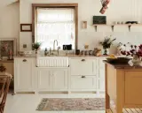 Urocza kuchnia retro inspiracje: białe szafki, ceramiczny zlew, mosiężna armatura, koronkowy zazdrostka i stylowy dywanik.