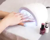 Dłonie z jasnoniebieskimi paznokciami pod lampą UV. W tle stoją buteleczki z lakierami. Lampa UV w ciąży - delikatna pielęgnacja.