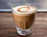 Ein Glas Cortado Kaffee mit Milchschaum-Latte-Art auf einem Holztisch.