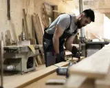 Carpintero ajustando una pieza de madera con una prensa. En el taller, se aprecian varios tablones y maquinaria, listos para crear acabados de madera perfectos.