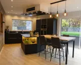 Cuisine noir et bois, mur gris texturé. Îlot central avec éclairage, table haute et tabourets. Fenêtre donnant sur jardin.