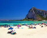 Spiaggia affollata con ombrelloni colorati, gente che si gode le vacanze al mare con bambini, acque turchesi e una montagna imponente sullo sfondo.