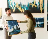 A man and woman admire a painting of blue trees at an art exhibition.