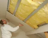 Pracownik montuje płyty gipsowo-kartonowe, kończąc ocieplenie dachu wełną mineralną.