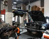 Un taller ordenado con un coche clásico negro con el capó abierto y otro azul en elevación. Motores y herramientas a la vista.