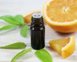 Flacon d'huile essentielle de citrus sinensis, entouré de feuilles vertes et de morceaux d'orange fraîche sur fond de bois clair.