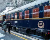 Ett par omfamnar varandra vid en snötäckt station, redo för en resa med Orientexpressen. Tågets mörkblå vagnar med gyllene detaljer vittnar om dess klassiska orientexpressen rutt.