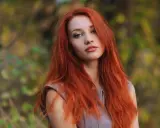Eine Frau mit leuchtend roten Haaren steht im Freien. Ihre langen, feurigen Haare umrahmen ihr Gesicht, während sie in die Kamera blickt. Die Natur im Hintergrund ist unscharf und zeigt herbstliche Farben.
