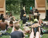 Un gruppo di persone rilassate su un prato in un **boutique festival**, con un DJ che suona musica in sottofondo.