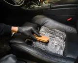 Mano enguantada cepilla asiento de cuero negro con espuma, mostrando cómo limpiar asientos de cuero para un coche impecable.