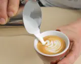 Barista gießt cremigen Milchschaum in eine Tasse Kaffee. Der Cappuccino Milchschaum bildet ein schönes Muster.