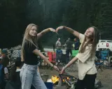 Dwie dziewczyny tworzą serce z dłoni na tle ogniska i lasu. To idealny okres na wakacjach, pełen radości i wspólnych chwil.