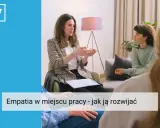 Kobieta w marynarce tłumaczy coś dłonią, siedząc naprzeciwko słuchaczy. Widać, że to rozmowa o empatii w pracy, która buduje lepsze relacje.
