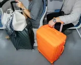 Dwie walizki, jedna pomarańczowa, druga ciemnozielona, obok siedzących osób w samolocie.