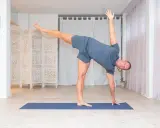 Man utför en dynamisk yoga-position på en blå matta i ett ljust rum med vita draperier och en vikbar rumsavdelare.