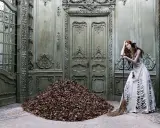 A woman in a tattered gown leans on a broom beside a mountain of leaves in a grand, ornate room, a scene captured by Eugenio Recuenco.