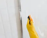 Ręka w żółtej rękawiczce czyści szklaną kabinę prysznicową gąbką. Dowiedz się, czym umyć kabinę prysznicową ze szkła, by lśniła czystością.