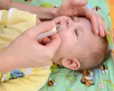 Mani che usano un aspiratore nasale per un neonato con raffreddore. Il piccolo indossa una tutina gialla con disegni di animali.