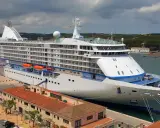 Grande nave da crociera attraccata nel porto Mahon, con edifici storici e colline verdi sullo sfondo.