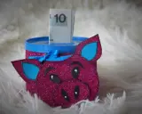 Urocza skarbonka diy w kształcie różowej świnki z brokatem, z niebieskimi uszami i kokardką. W środku widać banknot 10 zł.