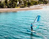 Mężczyzna na desce z żaglem pływa po turkusowej wodzie. Na brzegu widać plażę i drzewa.