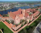 Malbork, największy zamek w Europie, rozciąga się wzdłuż rzeki, otoczony murami obronnymi i zielonymi dziedzińcami.