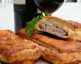 Pâté en croûte, une spécialité lorraine savoureuse, avec une garniture de viande visible. Une bouteille de vin rouge et du persil complètent la scène.