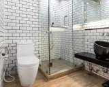 Baño moderno con azulejos blancos y negros. El suelo de madera se ve primero, luego los azulejos. Inodoro, ducha de cristal y lavabo negro.
