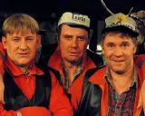 Tre män i orangea jackor och kepsar, en av dem med en uppstoppad älg i bakgrunden. Rollistan i Pistvakt.