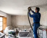 Hombre con mono azul lijando techo con gotelé para alisar paredes. Protege muebles y suelo con plásticos.