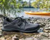 Szare buty sportowe na kamienistej plaży, idealne jako jakie buty na kajaki. W tle żółty kajak na wodzie.