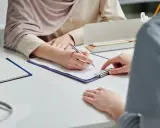 Dwie osoby przy biurku wypełniają dokumenty, jedna pisze, druga wskazuje miejsce.