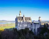Sagolikt slott i Tyskland, Neuschwanstein, reser sig majestätiskt på en klippa med en sjö och grönskande landskap i bakgrunden.