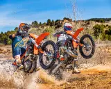 Dos pilotos de enduro compiten en un terreno embarrado, uno con motor 2t y otro 4t, salpicando agua.
