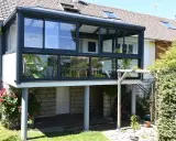 Une extension véranda toit plat moderne, avec de larges baies vitrées, s'intègre parfaitement à la maison. Un fauteuil vert est visible à l'intérieur.