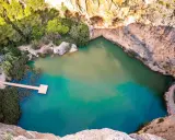 Un **charco azul** en Chiva, rodeado de rocas y vegetación. Un muelle de madera se adentra en sus aguas turquesas.