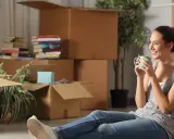 Une femme boit une tasse de café, entourée de nombreux cartons de déménagement.