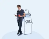 Młody mężczyzna z telefonem i napojem stoi obok kiosku do drukowania zdjęć z napisem "DRUKOWANIE...".