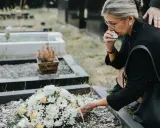 Kobieta w żałobie klęczy przy grobie, ocierając łzy. Czy zasiłek pogrzebowy należy się każdemu? Wieniec z białych i żółtych kwiatów leży na ziemi.