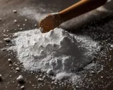 Stos białego proszku, być może soda oczyszczona na łupież pstry, z drewnianą łyżką w tle.