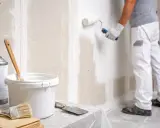 Un peintre applique une couche de peinture sur un mur en Fermacell. Matériel de peinture à proximité.