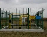Ogrodzony obiekt gazowy z żółtymi rurami. Tablice ostrzegawcze informują o zagrożeniu. Widać pola uprawne w tle, co sugeruje odległość budynku od gazociągu średniego ciśnienia.